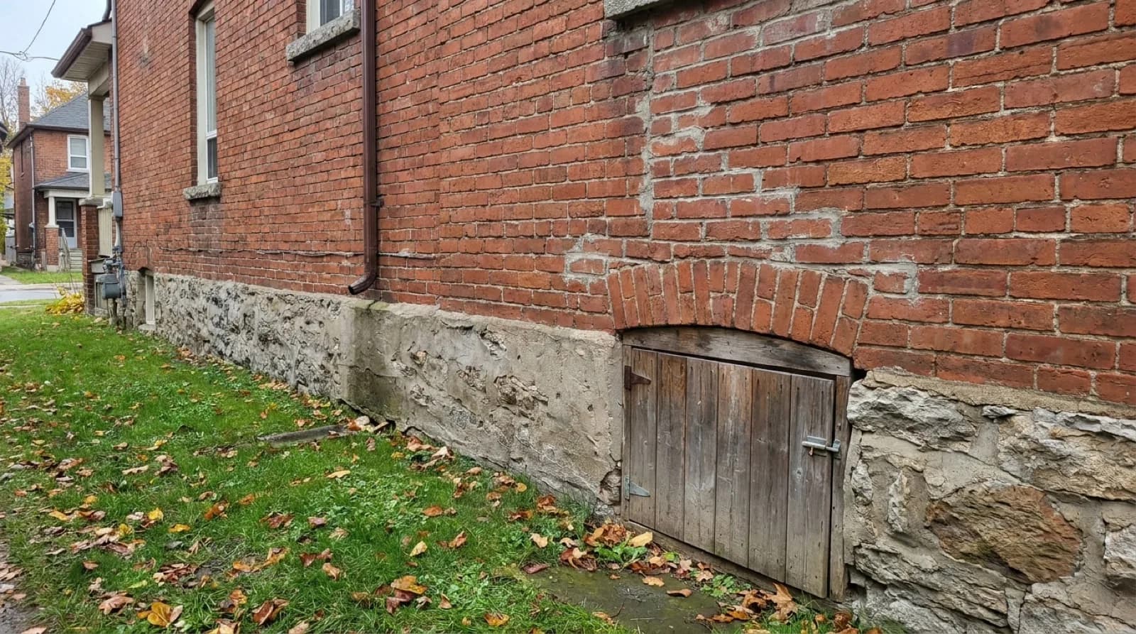The side wall of an older red brick house in Toronto, with a rough stone and concrete foundation and a small weathered wooden hatch under a brick arch near the ground, beside a grassy side yard with fallen leaves.