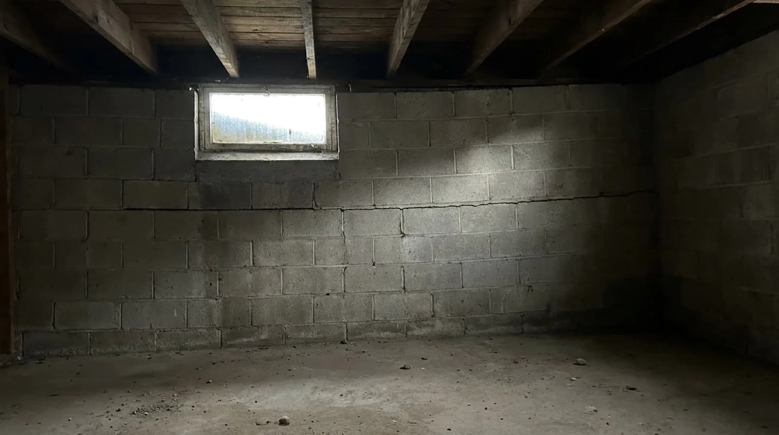 An empty unfinished basement in an older house, lit by one small window, where a grey concrete block foundation wall has a long horizontal crack running along a mortar joint below the window, under old wooden floor joists