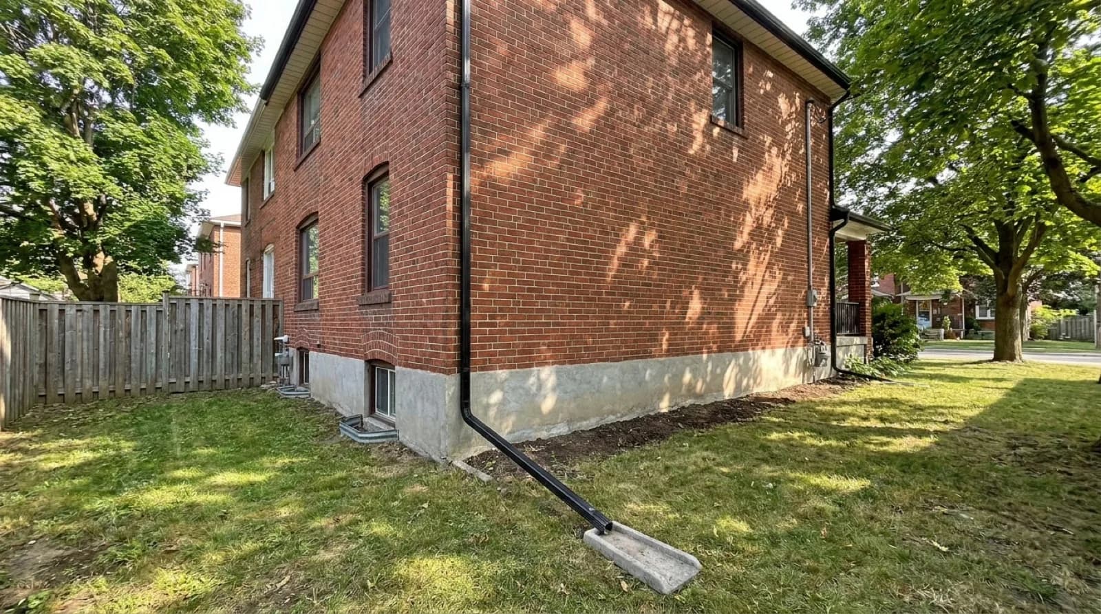 The side of an older red brick house on a leafy street, with a black downspout emptying onto a concrete splash pad on the lawn, a grey parged foundation along the base and a small basement window.